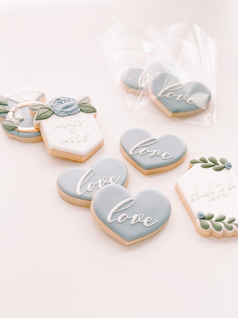 May include: Assortment of decorated sugar cookies with blue and white icing. The cookies feature the words "love", "bride to be", and "miss to mrs".