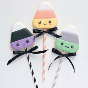 May include: Three Halloween-themed candy corn cookies on sticks, each with a different color scheme and a black bow. One is orange, yellow, and white, another is purple, black, and white, and the third is green, gray, and white.