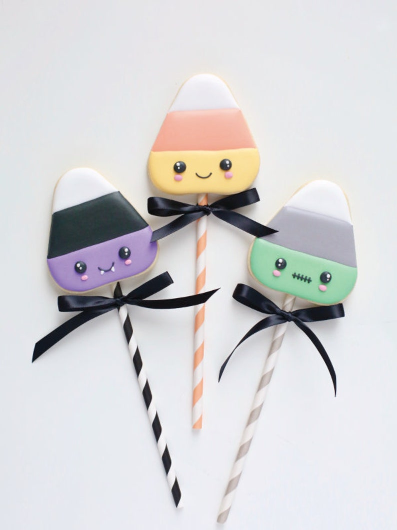 May include: Three Halloween-themed candy corn cookies on sticks, each with a different color scheme and a black bow. One is orange, yellow, and white, another is purple, black, and white, and the third is green, gray, and white.