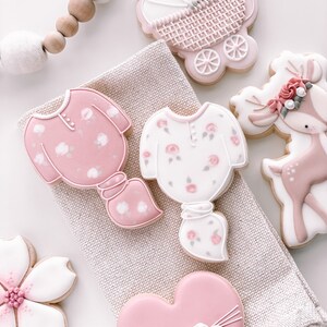 K&ouml;nnte beinhalten: Ein Set aus sechs rosafarbenen und wei&szlig;en, verzierten Zuckerkeksen. Die Kekse sind in Form eines Kinderwagens, eines Herzens, eines Rehs und zweier Strampler geformt. Die Kekse sind mit Blumen, Schleifen und anderen Details verziert.