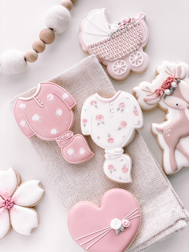 K&ouml;nnte beinhalten: Ein Set aus sechs rosafarbenen und wei&szlig;en, verzierten Zuckerkeksen. Die Kekse sind in Form eines Kinderwagens, eines Herzens, eines Rehs und zweier Strampler geformt. Die Kekse sind mit Blumen, Schleifen und anderen Details verziert.