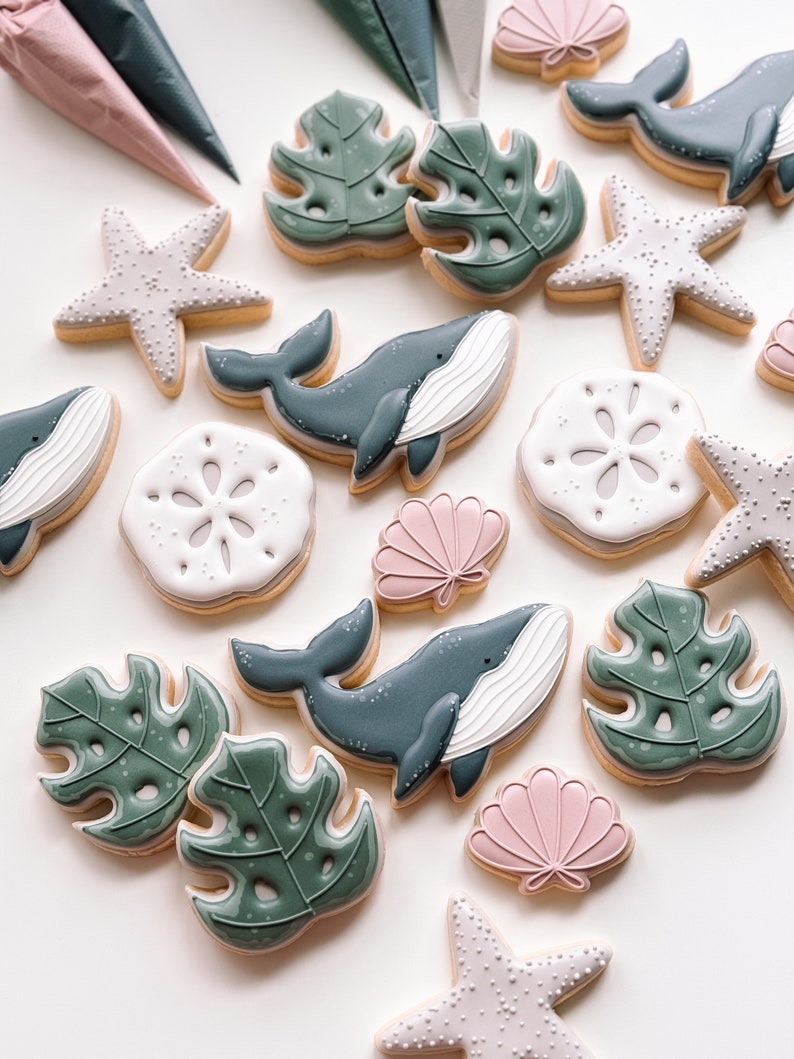 Op de afbeelding: Een verzameling gedecoreerde suikerkoekjes met verschillende ocean-thema ontwerpen, waaronder walvissen, schelpen, zeesterren en tropische bladeren. De koekjes zijn versierd met witte, roze en groene glazuur.
