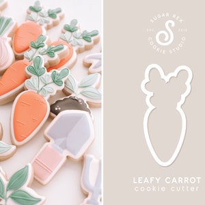 Puede incluir: Un cortador de galletas blanco en forma de zanahoria con hojas. El cortador est&aacute; sobre un fondo marr&oacute;n claro con el texto "Leafy Carrot cookie cutter" y un logotipo para Sugar Rex Cookie Studio.