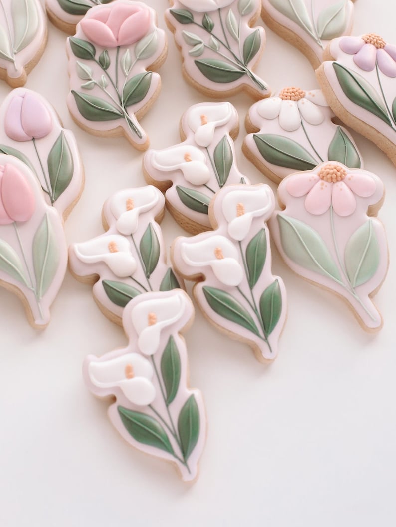 May include: A collection of decorated sugar cookies featuring various floral designs. The cookies are decorated with pink, white, and green icing, and each cookie features a different flower, including tulips, lilies, and daisies.