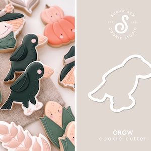 May include: A white crow cookie cutter with a simple design. The cutter is shown on a light brown background with the text "CROW cookie cutter" below it. The text "SUGAR REX COOKIE STUDIO EST 2019" is in the top right corner of the image.