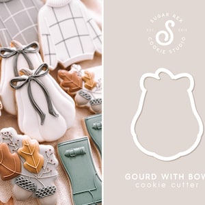 May include: A white cookie cutter shaped like a gourd with a bow on top. The cutter is on a light brown background with the text "Gourd with Bow cookie cutter" below it. The text "Sugar Rex Cookie Studio" is in a circle in the upper right corner of the image.