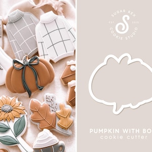 May include: A white cookie cutter in the shape of a pumpkin with a bow. The cookie cutter is on a light brown background with the text "PUMPKIN WITH BOW cookie cutter" below it.