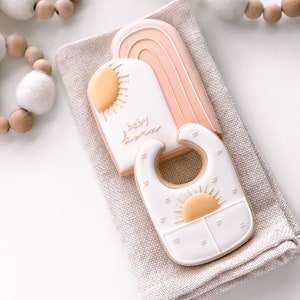 Puede incluir: Dos galletas de az&uacute;car decoradas con glaseado rosa pastel, blanco y amarillo. Una galleta tiene forma de arco&iacute;ris con las palabras "baby boom" escritas en ella. La otra galleta tiene forma de babero con un dise&ntilde;o de sol.