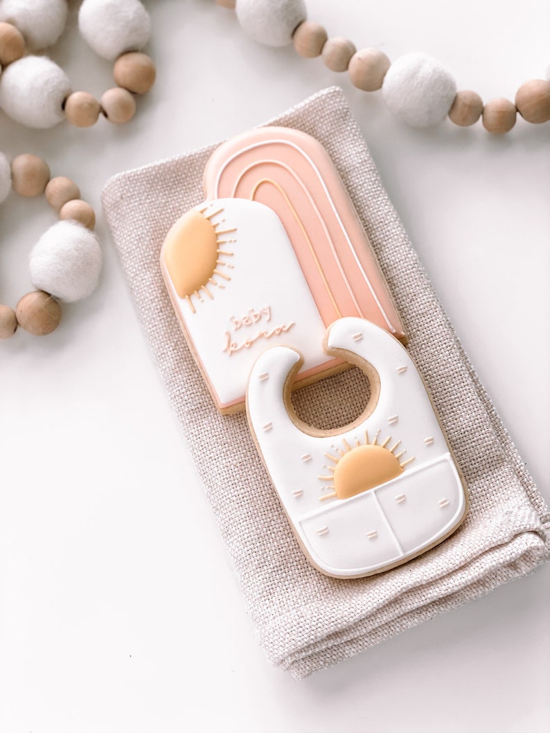 Puede incluir: Dos galletas de az&uacute;car decoradas con glaseado rosa pastel, blanco y amarillo. Una galleta tiene forma de arco&iacute;ris con las palabras "baby boom" escritas en ella. La otra galleta tiene forma de babero con un dise&ntilde;o de sol.