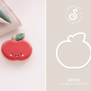 May include: A red apple-shaped cookie with a green leaf and a smiling face. The cookie is on a white surface, next to a beige cloth and green piping bags. A white apple cookie cutter is on a beige background with the text "APPLE cookie cutter".