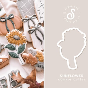 May include: A white cookie cutter in the shape of a sunflower. The cutter is on a light brown background with the text "SUNFLOWER cookie cutter" below it. The text "SUGAR REX COOKIE STUDIO EST. 2018" is in a circle above the cutter.