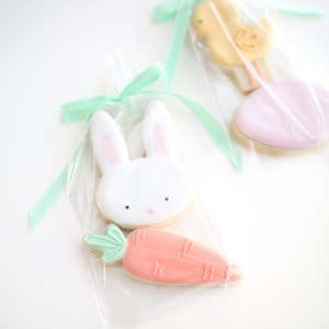 May include: Two cellophane bags tied with green ribbon, each containing a set of decorated sugar cookies. One bag contains a white bunny cookie and an orange carrot cookie. The other bag contains a yellow chick cookie and a pink heart cookie.