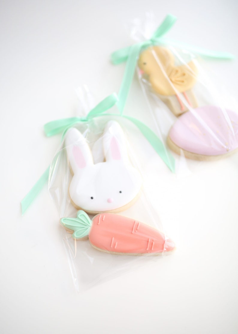 May include: Two cellophane bags tied with green ribbon, each containing a set of decorated sugar cookies. One bag contains a white bunny cookie and an orange carrot cookie. The other bag contains a yellow chick cookie and a pink heart cookie.