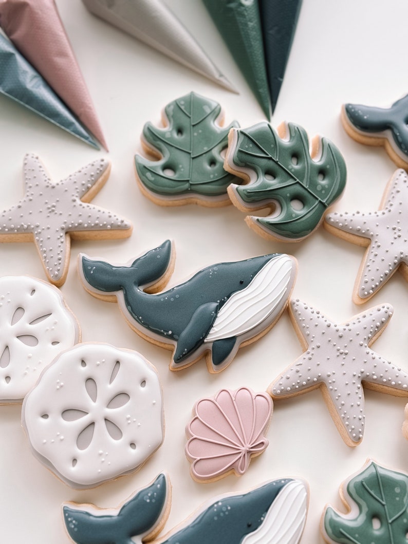May include: A collection of decorated sugar cookies featuring a variety of ocean-themed designs, including whales, seashells, starfish, and tropical leaves. The cookies are decorated with royal icing in shades of blue, green, pink, and white.