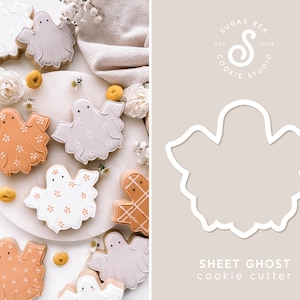 May include: A sheet ghost cookie cutter, a white plastic stencil, with a design of a ghost with a rounded head and a flowing body. The cutter is shown on a light brown background with the text "SHEET GHOST cookie cutter" below it.