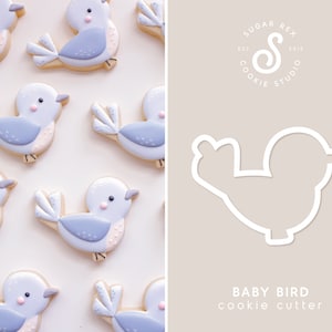 May include: A cookie cutter in the shape of a baby bird. The cutter is white and is shown against a light brown background. The text "BABY BIRD cookie cutter" is below the cutter.