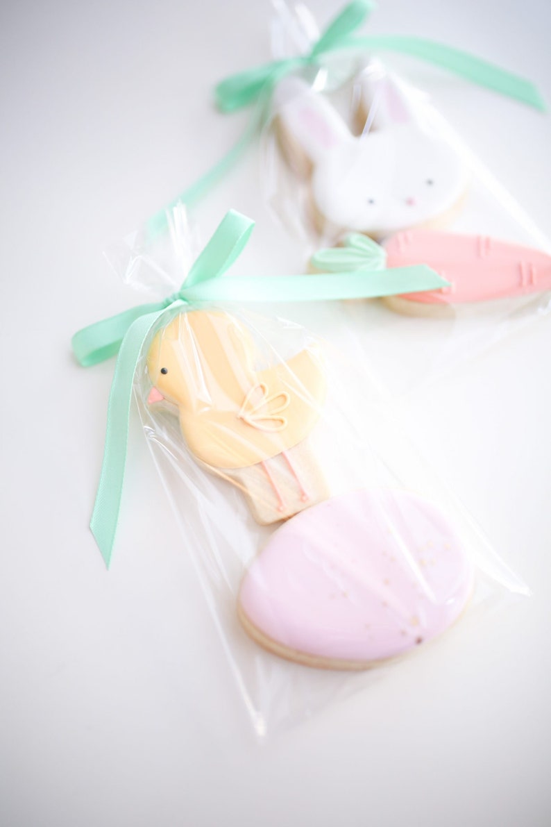 Op de afbeelding: Paaskoekjes in doorzichtige cellofaan zakjes vastgebonden met mintgroene linten. De koekjes bevatten een gele kuiken, een roze ei, een witte konijn en een wortel. De koekjes zijn versierd met pastelkleuren.
