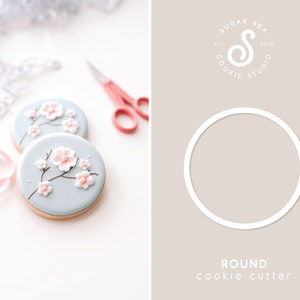 May include: Two round sugar cookies decorated with light blue icing and pink cherry blossoms. The cookies are on a white surface. A white circle with the text "ROUND cookie cutter" is on a light brown background.