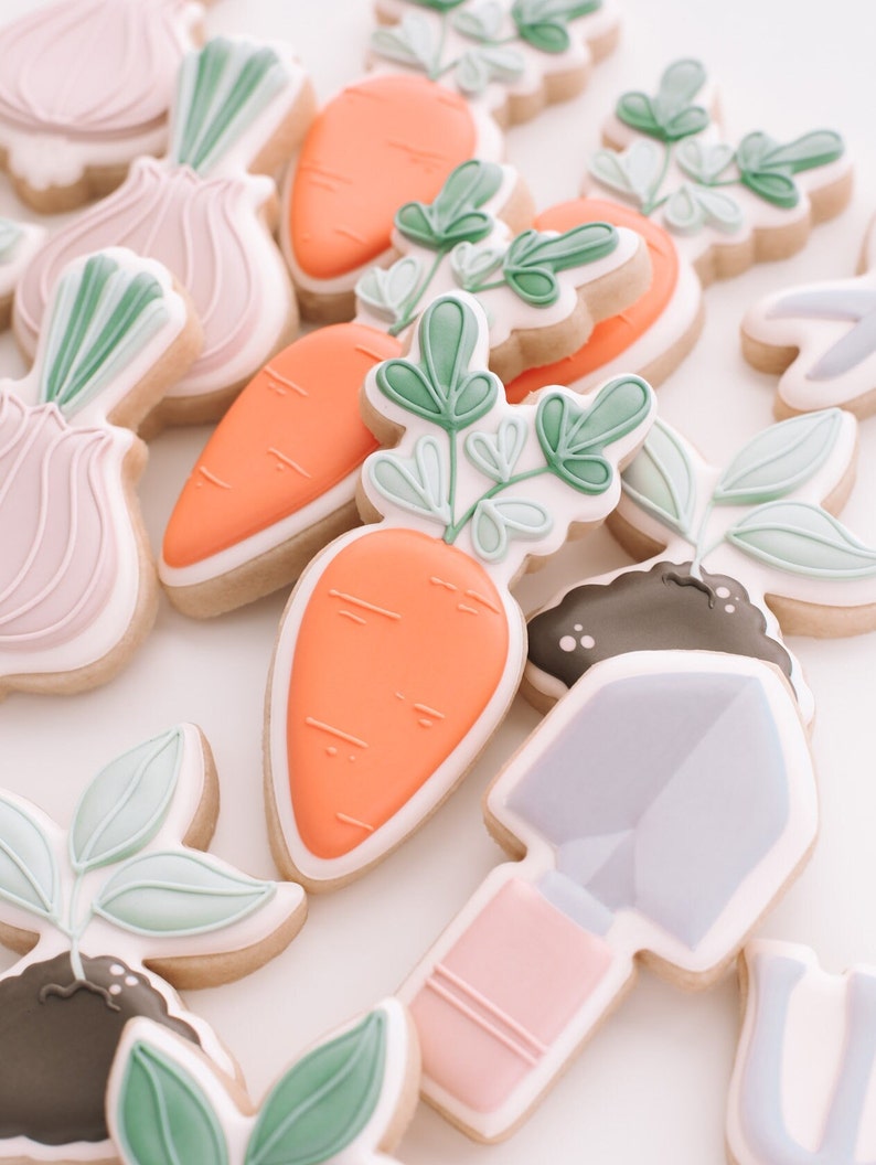 Puede incluir: Un conjunto de galletas de az&uacute;car decoradas con un tema de jard&iacute;n. Las galletas est&aacute;n decoradas con hojas verdes, zanahorias naranjas, cebollas rosas y una pala morada.
