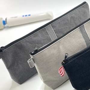 May include: Three zippered pouches made of different fabrics. The pouches are gray, beige, and black. Each pouch has a black zipper and a white and black striped ribbon. The black pouch has a red and white striped ribbon.