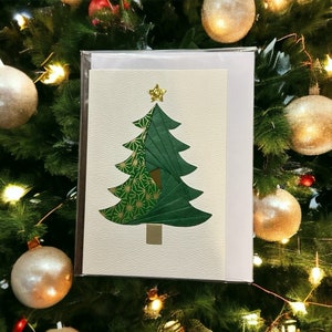May include: A Christmas card featuring a green origami Christmas tree with a gold star on top. The tree is made of folded paper with a patterned design on one side. The card is on a cream-colored background.
