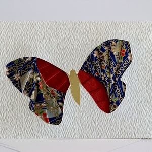 May include: A white card with a red, gold, and blue butterfly made of fabric. The butterfly is cut out of the card and the fabric is layered to create a 3D effect.