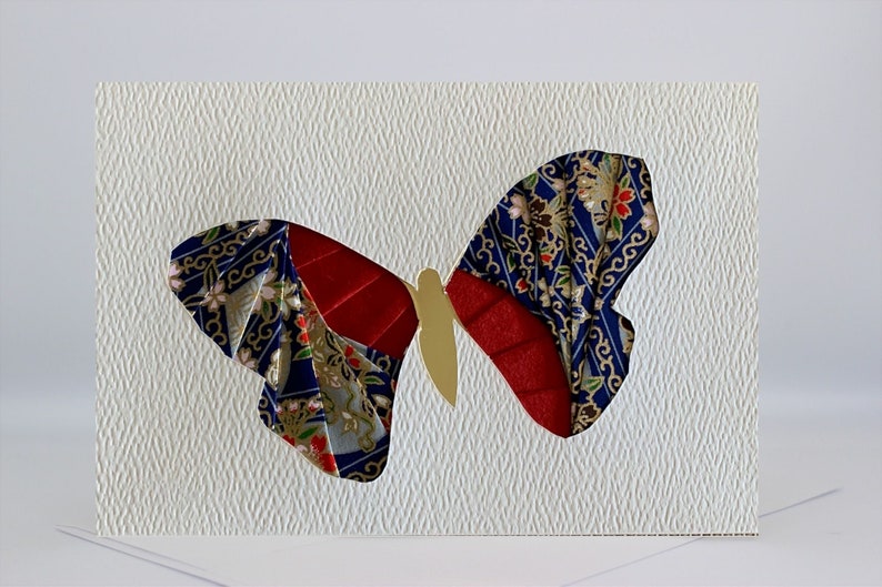 May include: A white card with a red, gold, and blue butterfly made of fabric. The butterfly is cut out of the card and the fabric is layered to create a 3D effect.