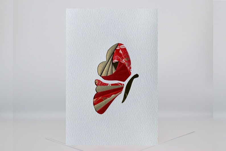 May include: A white card with a red and gold butterfly made of paper. The butterfly is cut out of the card and has a textured surface.