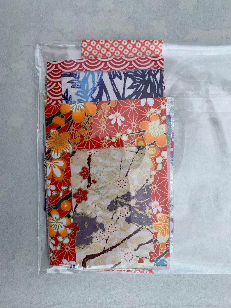May include: A clear plastic bag containing a stack of colorful patterned Japanese papers. The papers feature floral, geometric, and wave designs in shades of red, blue, gold, and white.