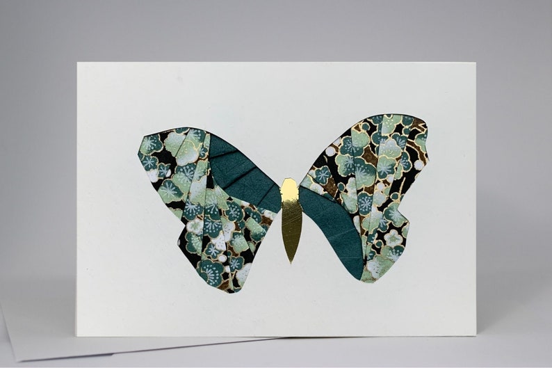 May include: A white card with a green and gold butterfly cut out of patterned paper. The butterfly has a floral design with black, white, and green colors.
