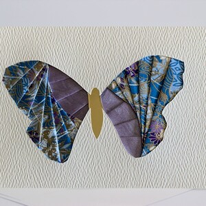May include: A handmade greeting card featuring a colorful butterfly made from fabric scraps. The butterfly is a light blue, purple, and gold color and is set against a white textured background.