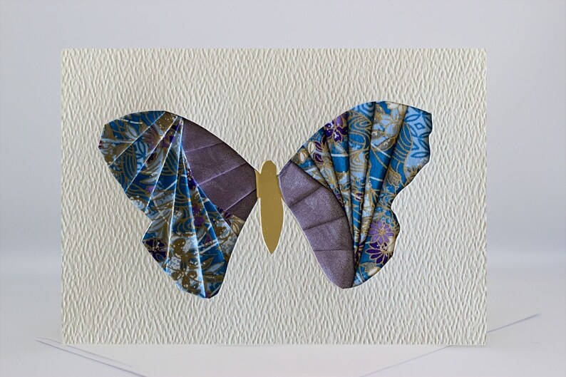 May include: A handmade greeting card featuring a colorful butterfly made from fabric scraps. The butterfly is a light blue, purple, and gold color and is set against a white textured background.