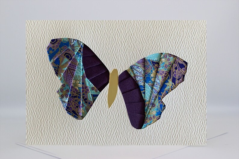 May include: A handmade greeting card featuring a purple and blue butterfly with gold accents. The butterfly is cut out of the card and layered with patterned paper.