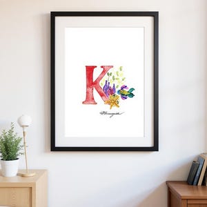 May include: Framed art print with a watercolour illustration of the letter "K" in red, decorated with marine life and foliage. The artwork includes a fish, starfish, and bottles, with the artist's signature at the bottom.