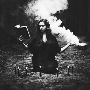 May include: A woman in a dark dress sits in a woodland setting, surrounded by candles and smoke. She holds a book in one hand and a cauldron in the other.