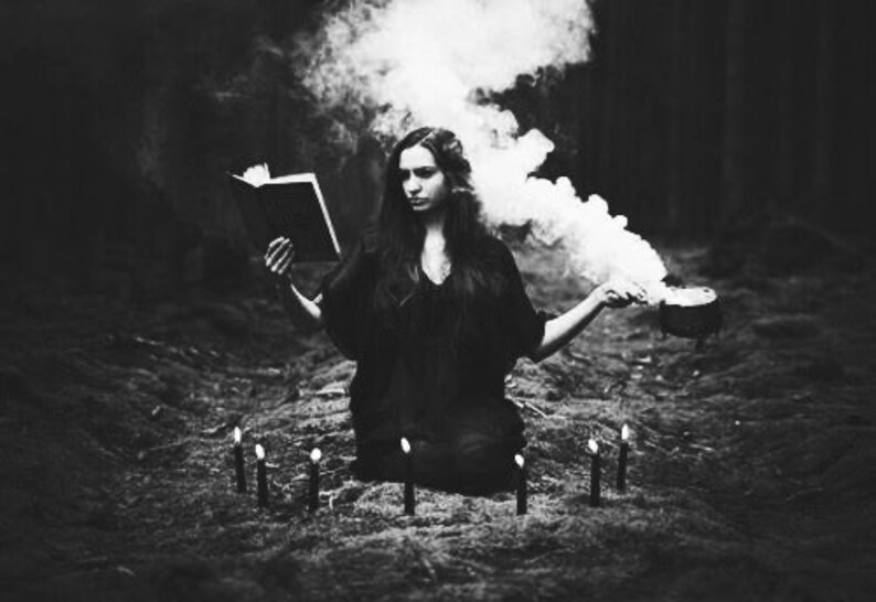 May include: A woman in a dark dress sits in a woodland setting, surrounded by candles and smoke. She holds a book in one hand and a cauldron in the other.