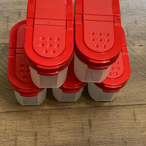 May include: A collection of red-lidded spice containers with clear, rectangular bodies. The lids have a flip-top design with small holes for dispensing. The containers are on a wooden surface.