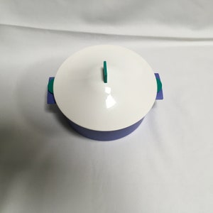 Puede incluir: Una olla arrocera de color morado y turquesa con una tapa blanca y un asa turquesa.