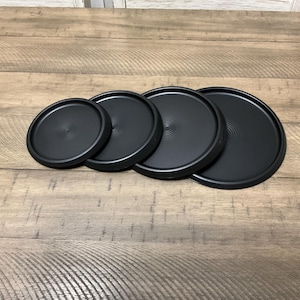 May include: Four black round coasters of varying sizes. The coasters are made of a smooth, hard material and have a slightly raised edge.