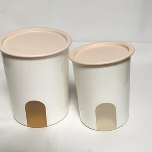 Op de afbeelding: Twee witte plastic kannen met beige deksels en een doorschijnend bruin accent. De kannen zijn van verschillende grootte.