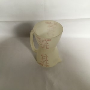 以下が含まれることがあります： 透明なオフホワイトのプラスチック製計量カップ。赤い目盛りでオンスとカップの量を表示。ユニークな砂時計型で、上部と下部が広くなっています。
