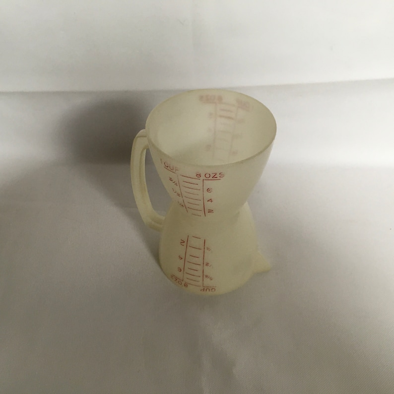 以下が含まれることがあります： 透明なオフホワイトのプラスチック製計量カップ。赤い目盛りでオンスとカップの量を表示。ユニークな砂時計型で、上部と下部が広くなっています。