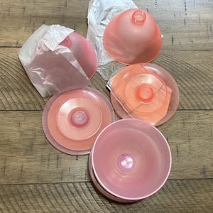 May include: A collection of pink plastic kitchen items, including a large bowl, several lids, and a funnel-shaped piece. The items are arranged on a wooden surface, with some still wrapped in white paper. The colors range from light to medium pink.