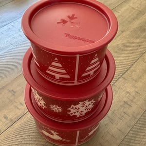 May include: Three red Tupperware containers stacked, each with a lid. The top lid has the Tupperware logo. The containers feature white festive designs: Christmas trees, snowflakes, and reindeer. Ideal for food storage.
