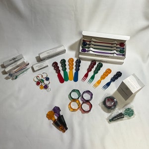 Peut inclure: Une collection de pièces de service en plastique multi-facettes et colorées, comprenant des fourchettes, des cuillères et un bouchon de bouteille. Les pièces sont disposées sur une surface blanche avec plusieurs boîtes portant les mentions "Simply Elegant" et "Tupperware".