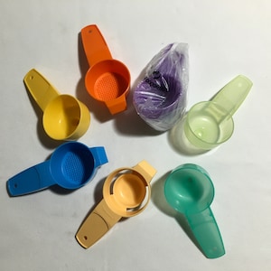 May include: A set of six colorful plastic measuring cups with different designs. The cups are yellow, orange, blue, green, light green, and purple. Some of the cups have a strainer design.