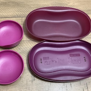 May include: A set of four magenta-colored plastic food containers. The set includes two oval-shaped containers, one with a lid, and two smaller round bowls. The containers are made of a durable plastic material.
