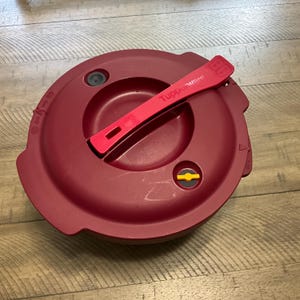 May include: Red plastic pressure cooker lid with a red handle that says "Tupperware" on it. The lid has a yellow valve and a small black knob.