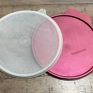 May include: Two Tupperware lids, one clear and one pink, rest on a wooden surface. The clear lid has the word "Tupperware" printed on it. The pink lid has a rounded handle and the word "Tupperware" printed on it.
