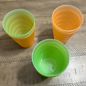 May include: Three plastic cups in shades of orange and green. The cups have a ridged design and are arranged on a wooden surface. The cups are empty and appear to be clean.